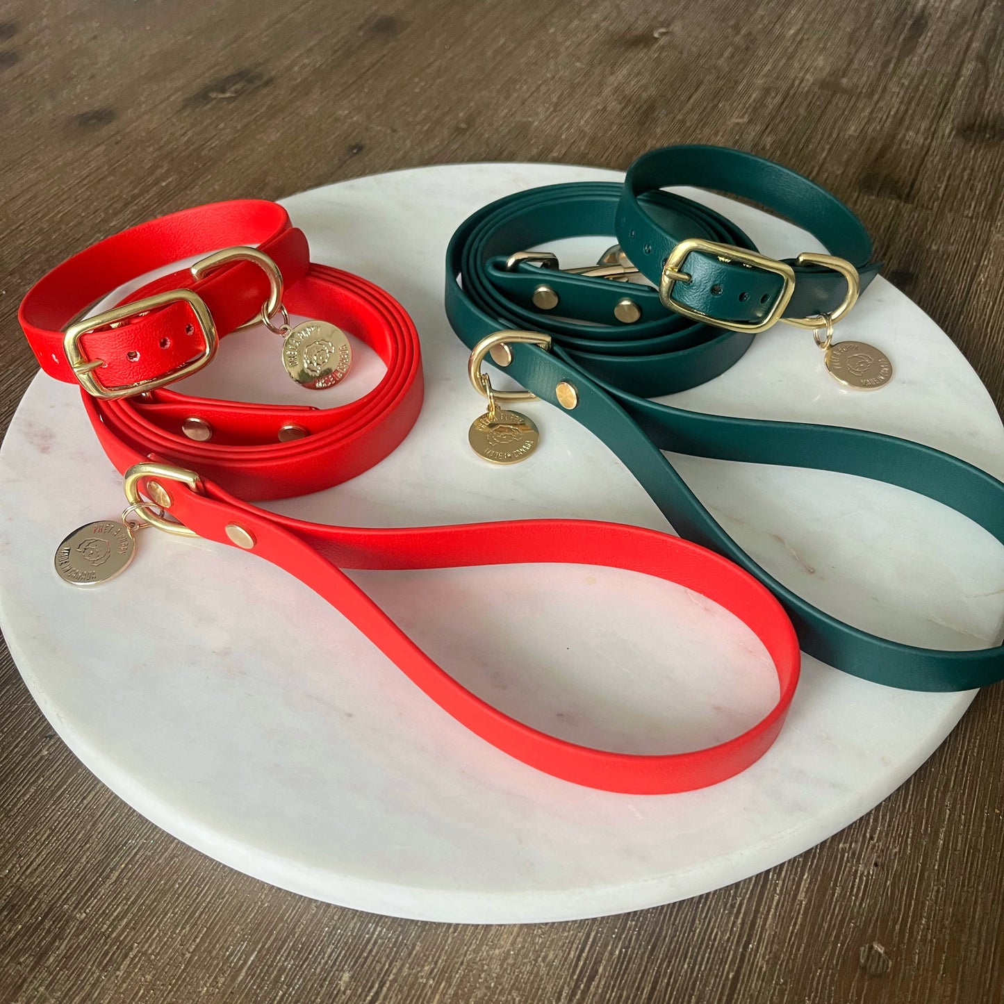 A forest green dog collar made from soft-to-touch, waterproof vegan leather, featuring shiny brass gold hardware including a buckle and D-ring. Attached to the collar is a small charm that adds a touch of elegance. The overall design is simple yet stylish, perfect for a fashionable pet. Also shown with a red collar